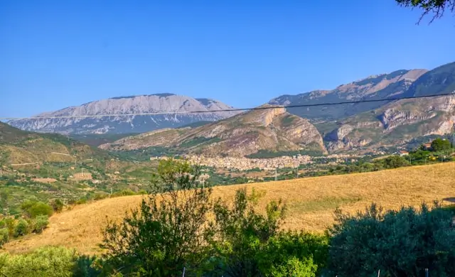 Panoramic view of Parco Delle Madonie, featuring lush green hills, rugged mountain peaks, and scattered trees under a clear blue sky.